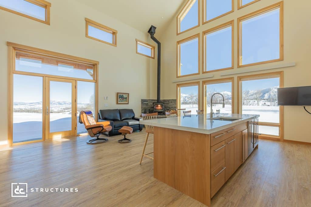 A modern living area with large windows, wood floors, island kitchen, black recliners, wood stove, and snowy mountain views.