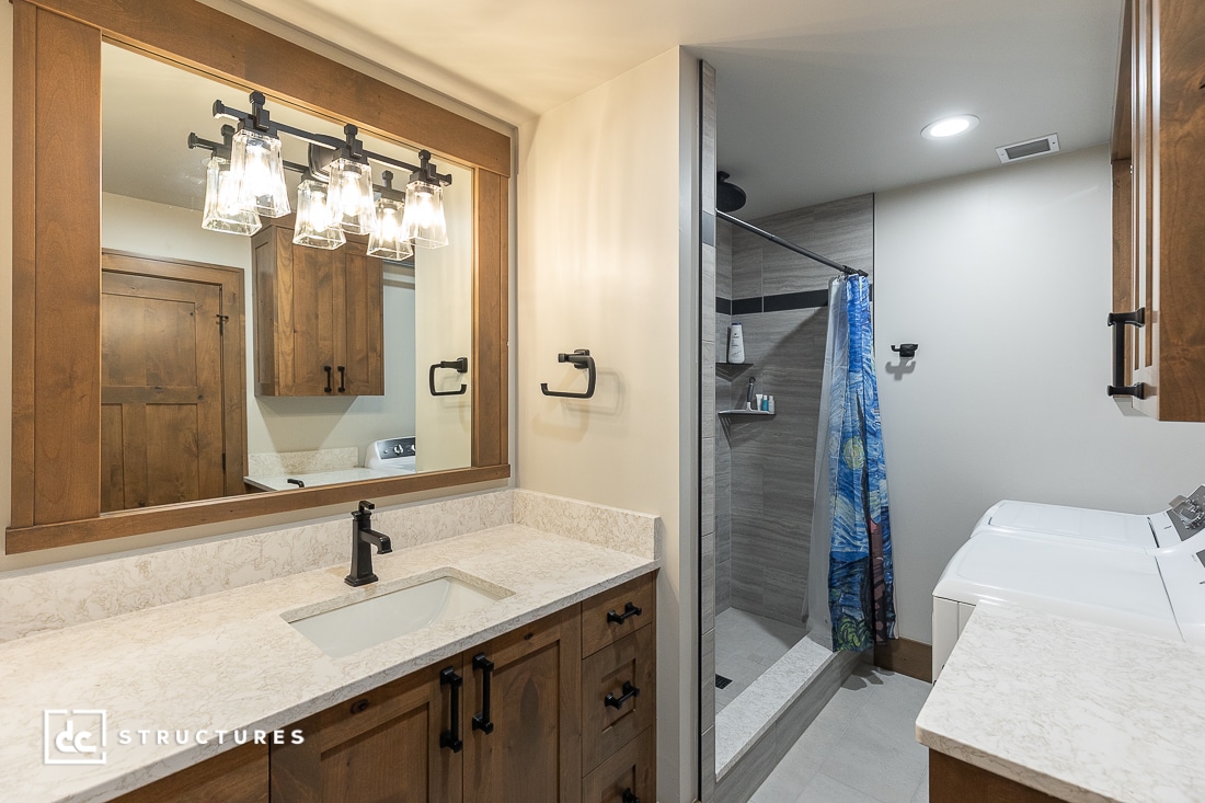 A modern bathroom and laundry space with wood cabinets, a large mirror, black fixtures, blue shower curtain, washer and dryer.