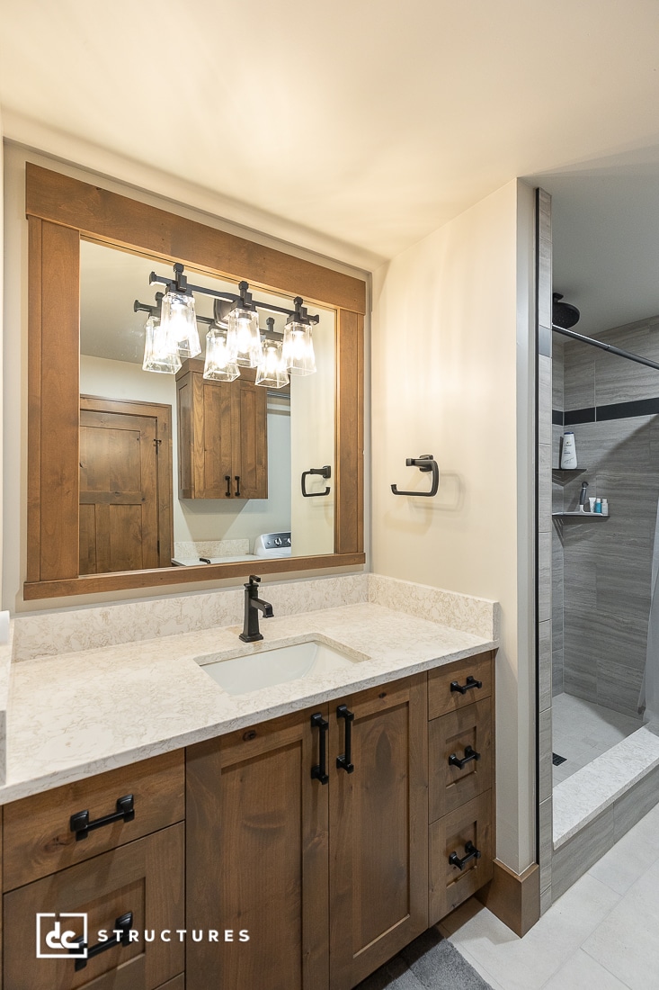 A modern bathroom with a wooden vanity, white countertop, rectangular sink, large mirror, black fixtures, and glass walk-in shower.