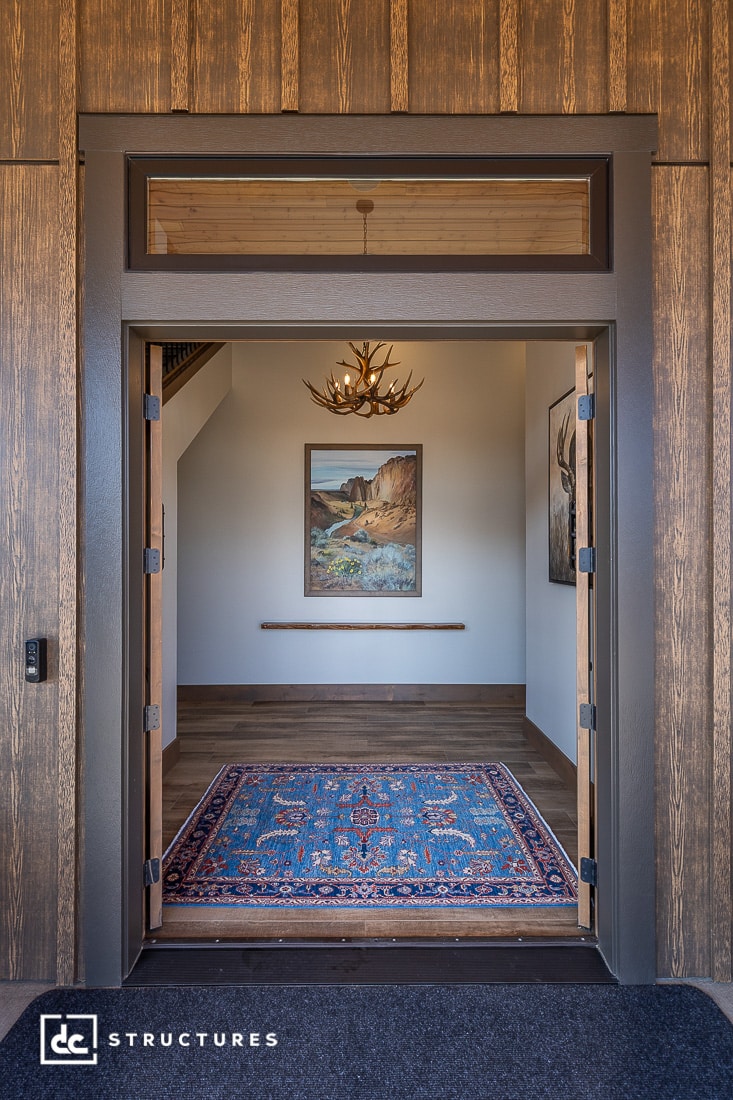 A view through open double doors into a foyer with a blue patterned rug, an antler chandelier, landscape painting, and wood-paneled walls.