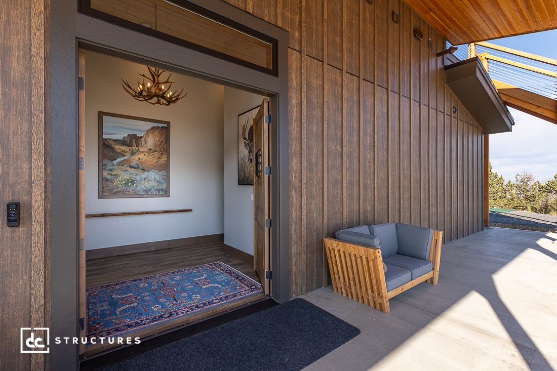 A modern wooden entryway with a blue-patterned rug, antler chandelier, and landscape painting. Outside, a wooden armchair with gray cushions sits on a concrete porch under a slanted roof.