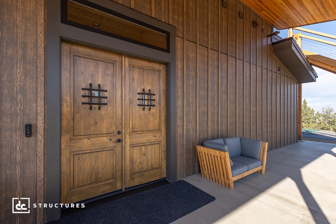 A modern wooden double front door with decorative iron grilles set in a wood-paneled exterior. A gray cushioned wooden bench sits to the right on a concrete porch.