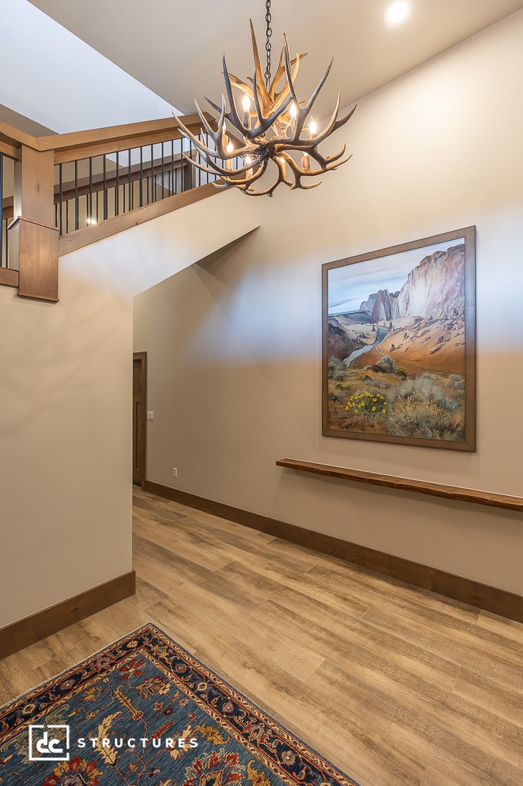A hallway with wood flooring, a colorful rug, a large landscape painting on the wall, an antler chandelier, and wooden railings above. The space feels warm and inviting.