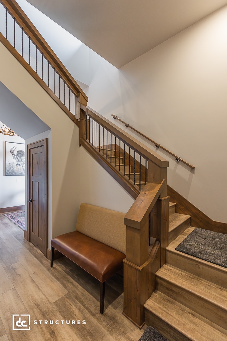 A wooden staircase with a beige cushioned bench beneath it, set against a light wall, wood flooring, closet door, and hallway art.
