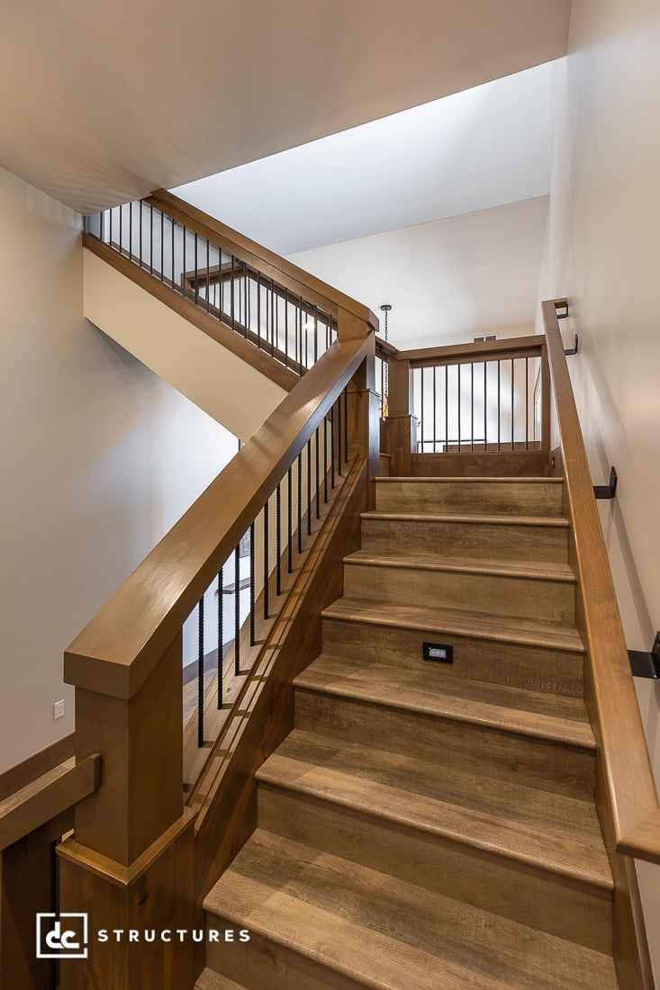 Interior view of a modern wooden staircase with metal railings, leading to an upper floor in a well-lit space. Walls are light-colored and clean.