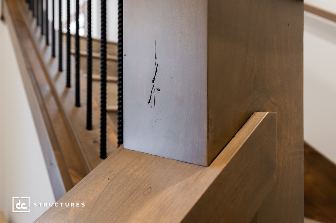 Close-up of a wooden staircase post with a visible scratch or crack on its surface; black balusters and stairs are seen in the background.
