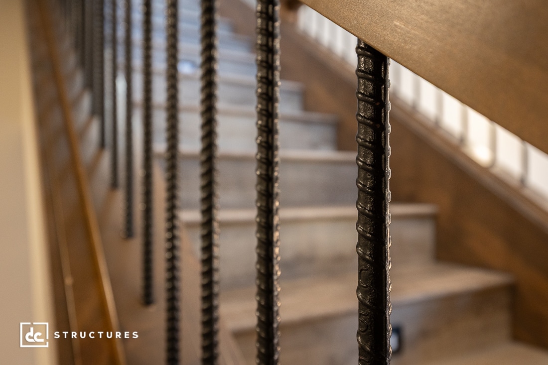 Close-up view of black metal stair balusters and a wooden handrail, with blurred concrete steps in the background.