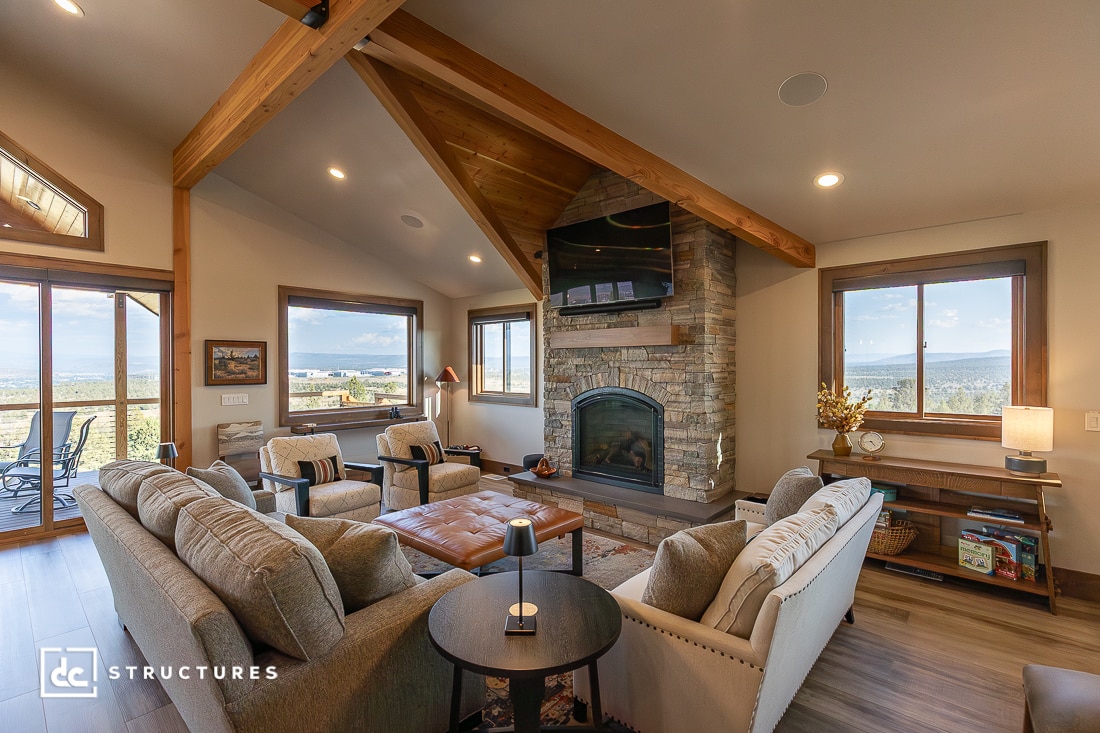 A cozy living room with neutral-toned sofas, a stone fireplace with a TV above, large windows, wood beam ceiling, modern and rustic decor.