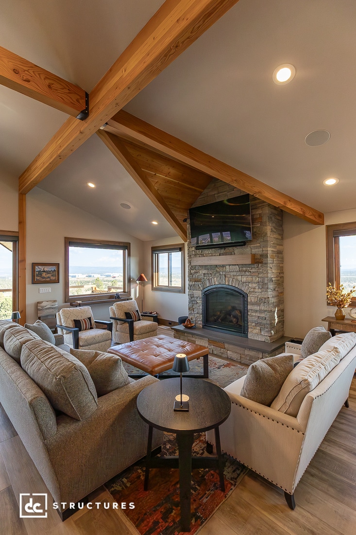 A cozy living room with wood beams, large windows, gray sofas, a stone fireplace, and a round side table with a lamp.