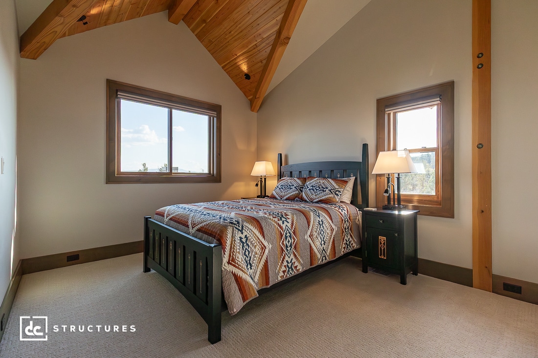A cozy bedroom with a high wooden ceiling, large windows, and a dark wooden bed with a patterned blanket. Two bedside tables with lamps flank the bed. Natural light fills the room.