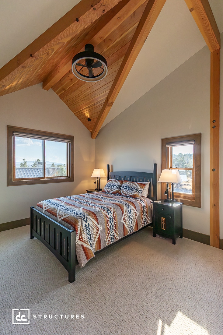 A cozy bedroom with a high wooden vaulted ceiling, dark wood bed with geometric bedding, matching nightstands with lamps, and two windows.