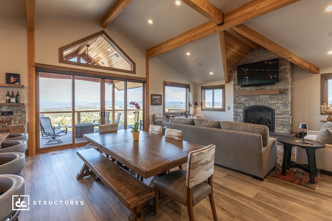 A spacious, modern living and dining area with wood beams, stone fireplace, large windows, wooden table, neutral furniture, and natural light.