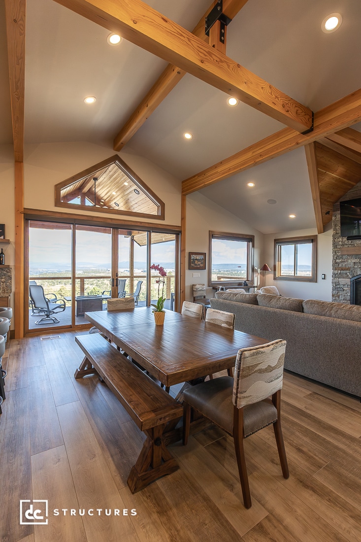 A spacious, modern living and dining area with wooden beams, large windows, a rustic table, and comfy seating overlooking the patio.