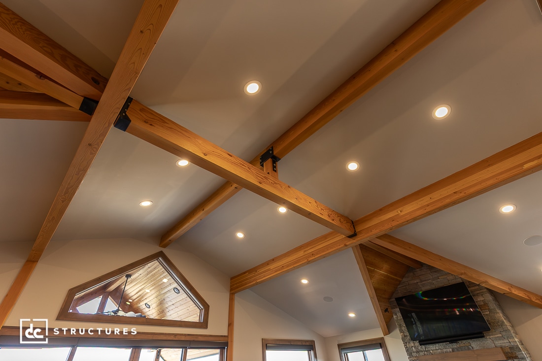 Exposed wooden beams and recessed lights on a vaulted ceiling, with part of a stone fireplace, TV, and windows visible below.