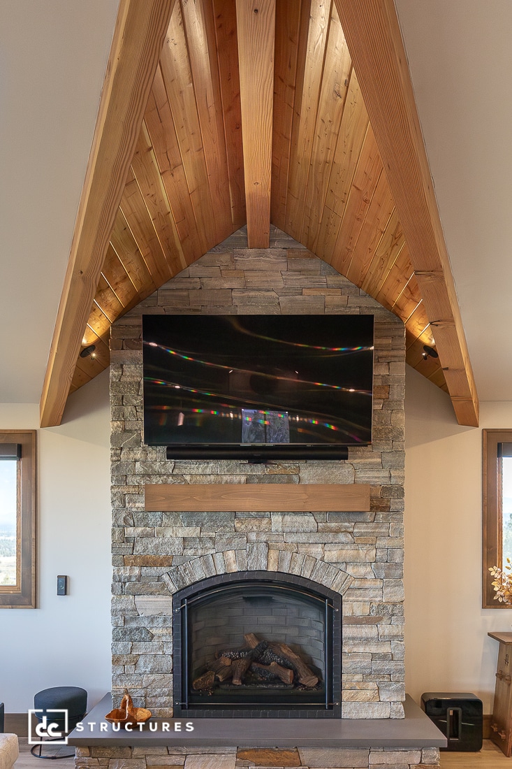 A vaulted ceiling with exposed wood beams, a stone fireplace, wooden mantel, and large flat-screen TV above the fireplace in a cozy room.