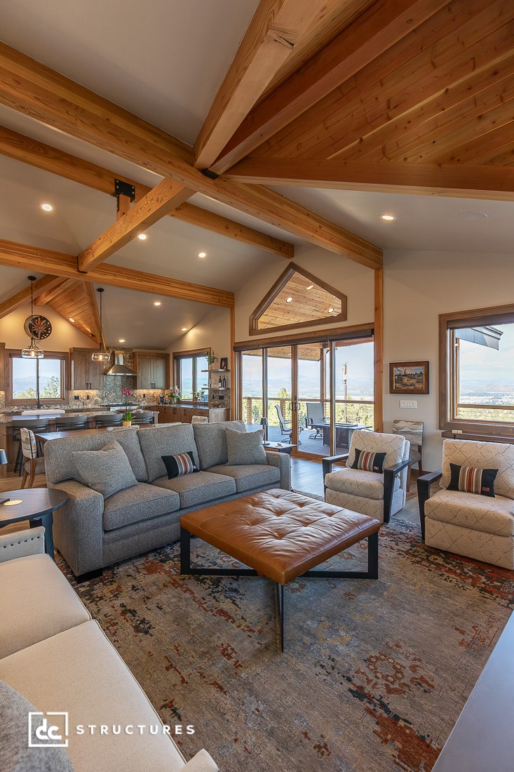 Spacious living room with exposed wooden beams, large windows with scenic views, gray sectional sofa, patterned chairs, brown tufted coffee table, and a patterned rug.