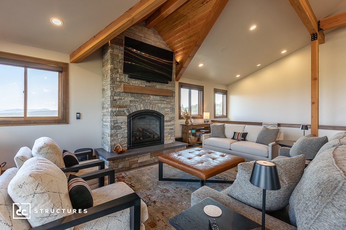 A cozy living room with vaulted wood ceilings, stone fireplace with a TV, neutral sofas, armchairs, leather ottoman, large windows.