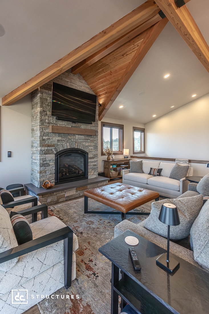 A modern living room with vaulted wooden beam ceiling, stone fireplace, wall-mounted TV, sofas, leather ottoman, and windows.
