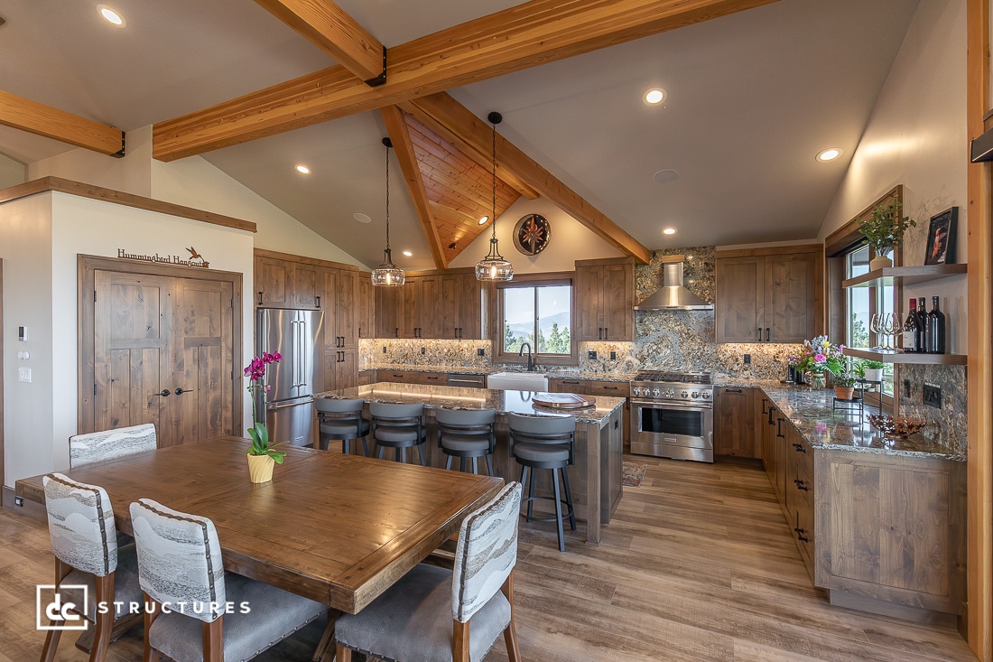 Modern kitchen and dining area with wooden beams, island and barstools, built-in appliances, stone backsplash, and natural light.