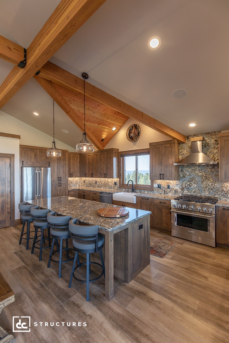 A rustic kitchen with wood beams, stone countertops, stainless steel appliances, a large island with blue chairs, and pendant lights.