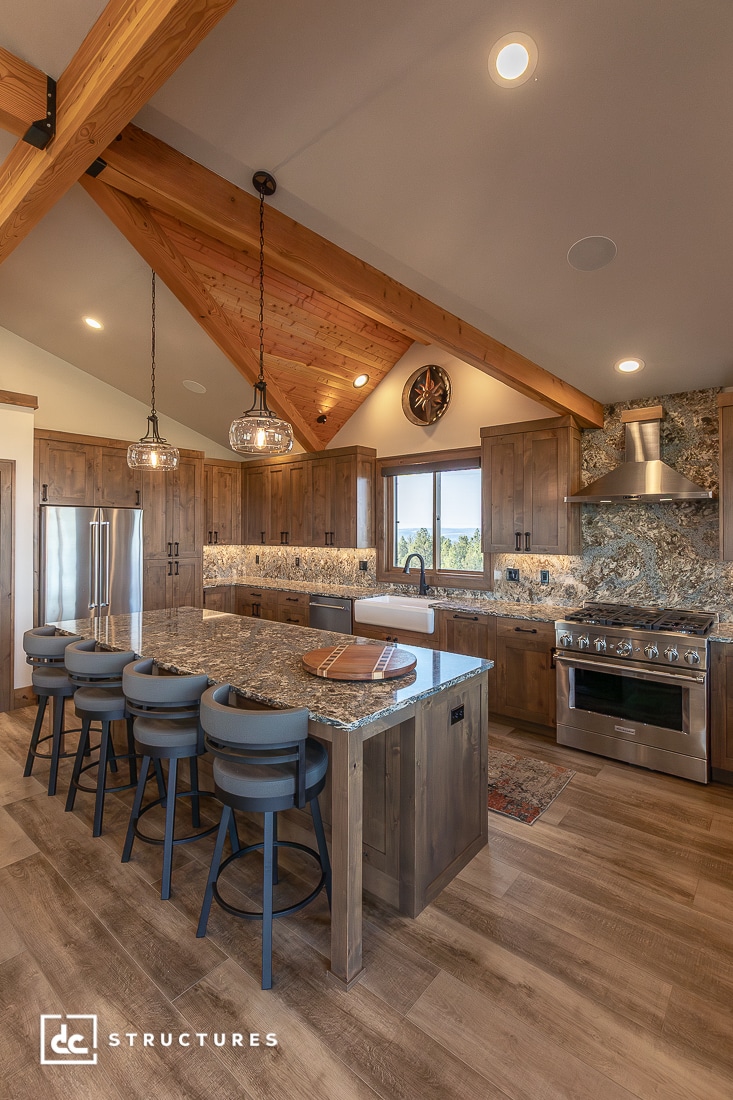 A modern rustic kitchen with wood cabinets, granite countertops, a large island with four gray barstools, stainless steel appliances, pendant lights, and exposed wooden beams on a vaulted ceiling.
