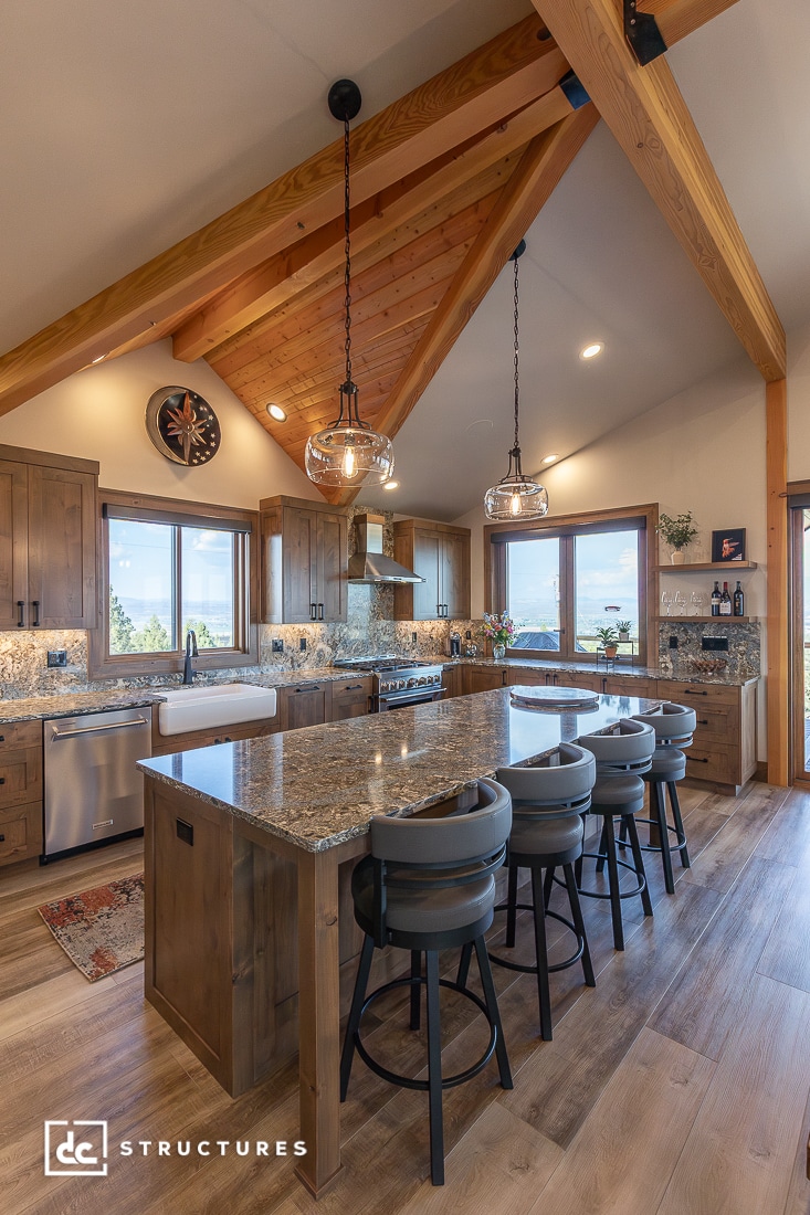 Modern rustic kitchen with wood beams, vaulted ceiling, large granite island, gray barstools, pendant lights, and mountain views.