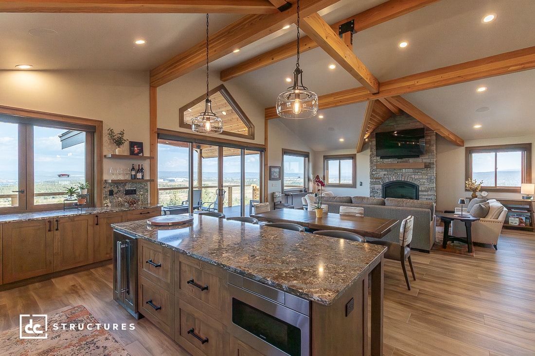 A spacious open-concept kitchen and living room with wood beams, granite countertops, large windows, wood cabinets, and a stone fireplace.