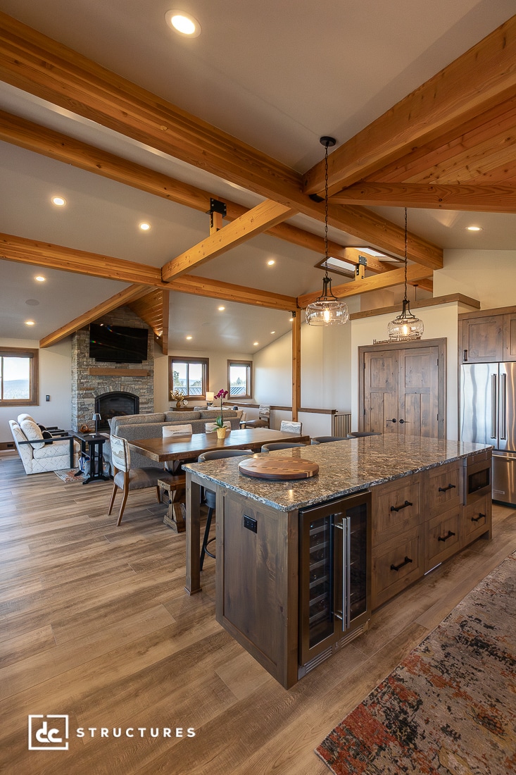 Open-concept kitchen and living area with exposed wooden beams, granite island, modern appliances, dining table, light fixtures, and a stone fireplace. Warm lighting and natural wood accents create a cozy atmosphere.