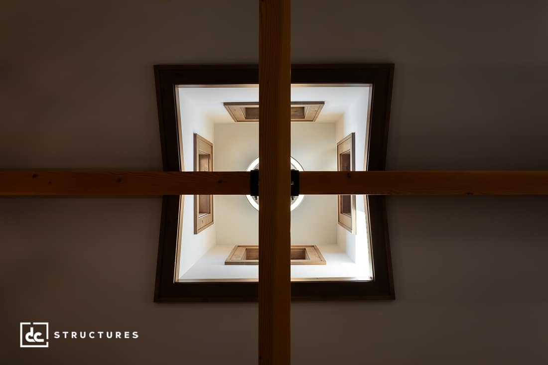 View looking up at a wooden beam cross with a square ceiling opening above, bordered by wood trim and featuring four small windows.