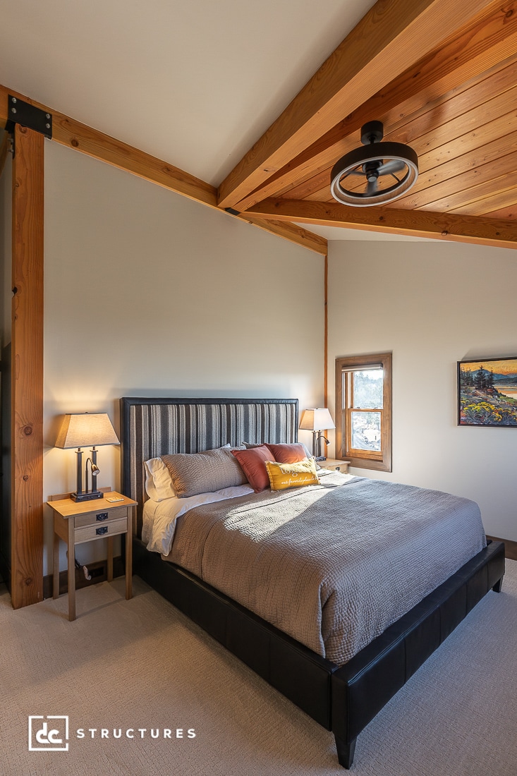 A modern bedroom with a large bed, striped headboard, bedside tables with lamps, wooden beams, a small window, and landscape painting.