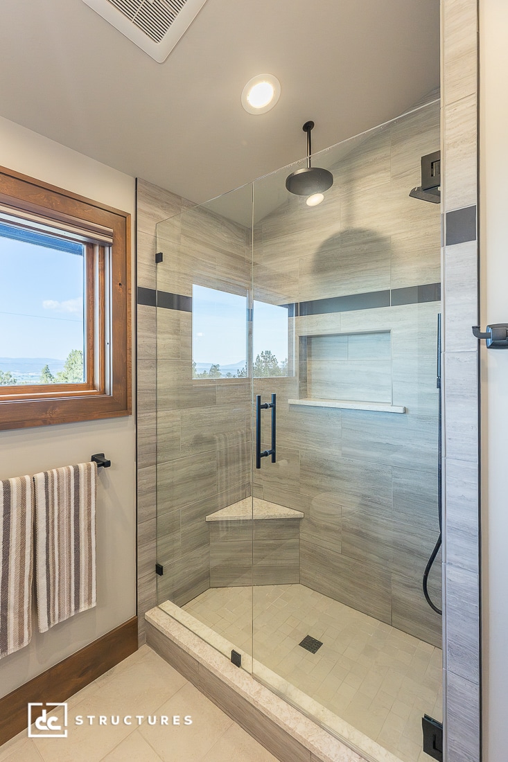 Modern glass-enclosed shower with beige tile walls, built-in bench, black rainfall showerhead, wood-framed window, and two striped towels.