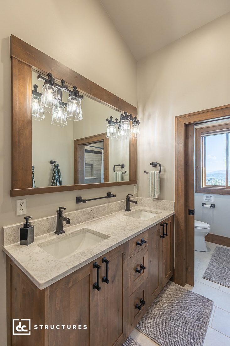 A modern bathroom with a double sink vanity, marble countertop, wooden cabinets, large framed mirror, decorative light fixtures, and a view into a toilet area with a window.