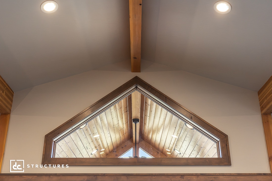 Interior view of a vaulted ceiling with exposed wooden beam and triangular window overlooking a wooden loft. Warm lighting and wooden accents create a cozy, inviting atmosphere.