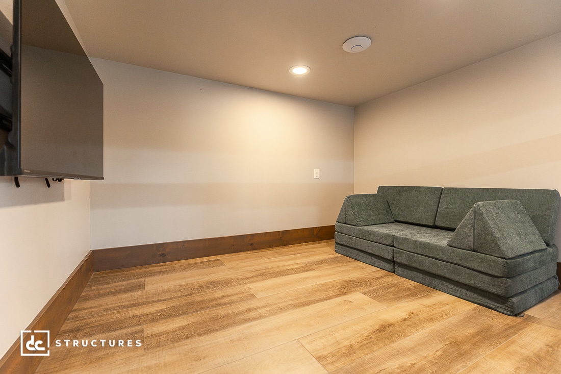 A simple room with light wood flooring, beige walls, a green modular sofa, and a wall-mounted flat-screen TV. The space is sparsely furnished with recessed ceiling lighting.