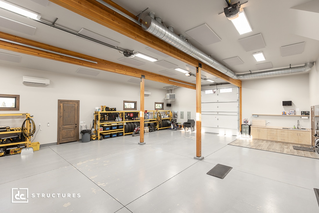 A spacious, well-lit garage with exposed wooden beams, organized shelves, a workbench, large garage doors, and a clean light gray floor.