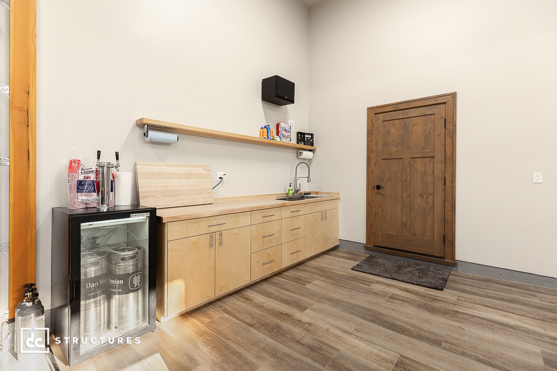 A modern kitchenette with light wood cabinets, a small sink, a mini fridge with two large drink kegs, a wooden door, wall shelf with drinks, and wood-look flooring.