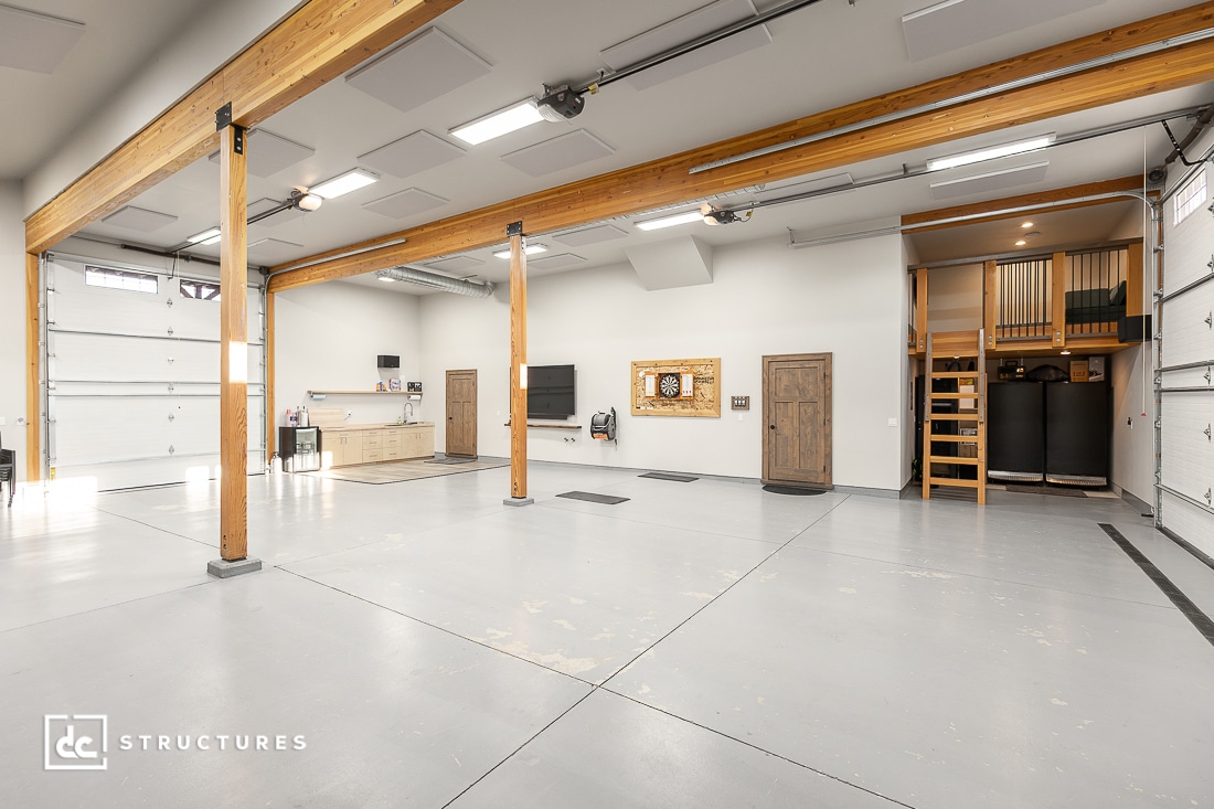 Spacious, clean garage with high ceilings, exposed wooden beams, white walls, and multiple garage doors. Kitchenette and wall-mounted TV. Loft area with stairs and water tanks underneath.