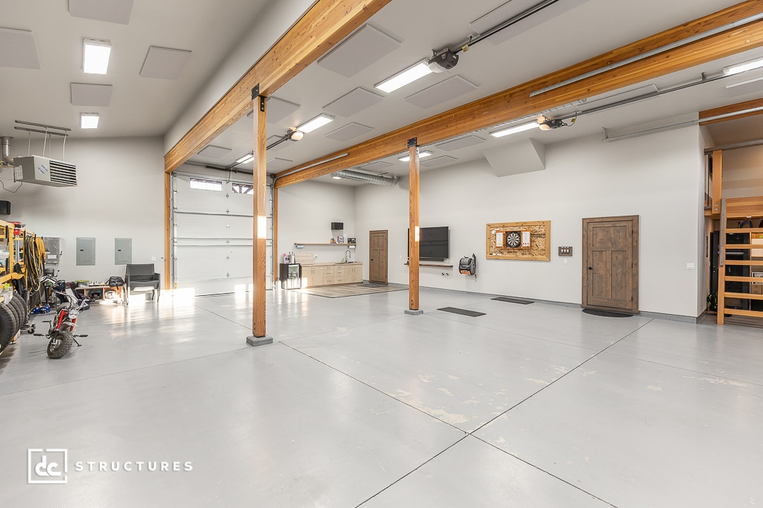 Spacious, modern garage with high ceilings, exposed wooden beams, and large windows. Workbenches, motorcycles, and tools line the walls. Clean light gray floor.