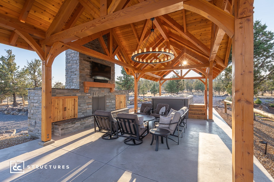 A spacious outdoor pavilion with a wooden gabled roof, stone fireplace, lounge seating, a round table, and scenic views of trees.