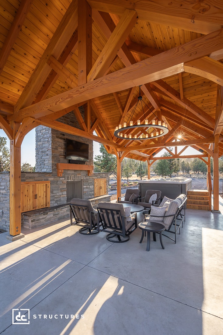 A spacious outdoor pavilion with exposed wooden beams, a stone fireplace, comfy patio seating, and a large circular chandelier.