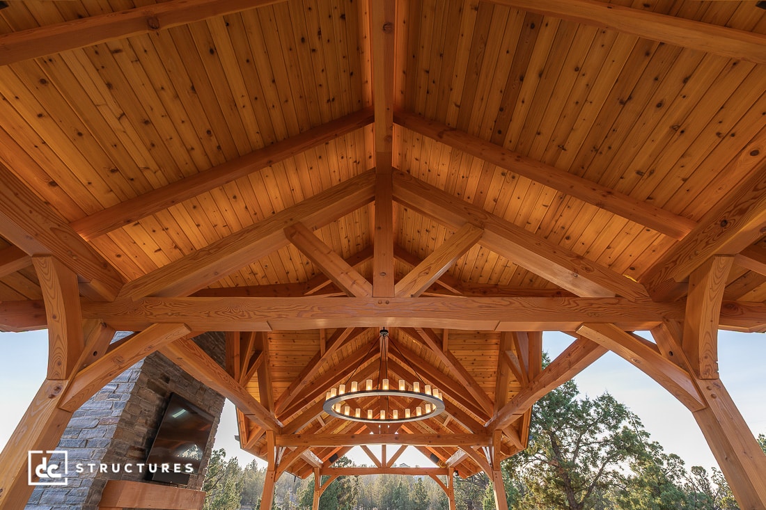 View of a wooden pavilion ceiling with exposed beams and a circular chandelier, showing intricate craftsmanship. Trees and stone fireplace are visible in the background.