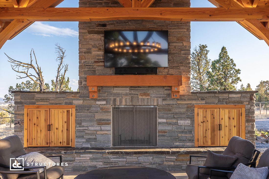 A stone outdoor fireplace with a TV above, flanked by wooden cabinets, sits under a pergola. Outdoor seating and trees behind.