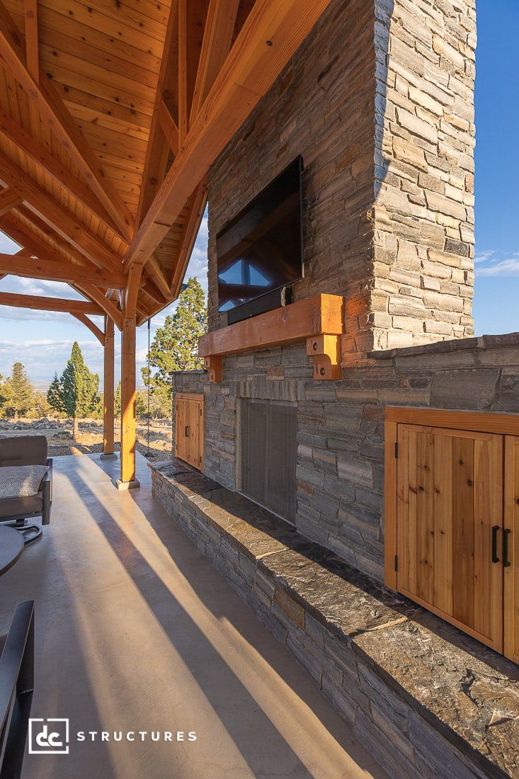 An outdoor patio has a stone fireplace with a TV above a wooden mantel, wooden cabinets, and pergola beams, with mountain views.