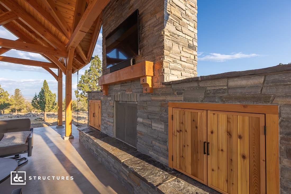 A covered outdoor patio with a stone fireplace, mounted TV, wooden storage cabinets, wooden beams, and trees in the background.