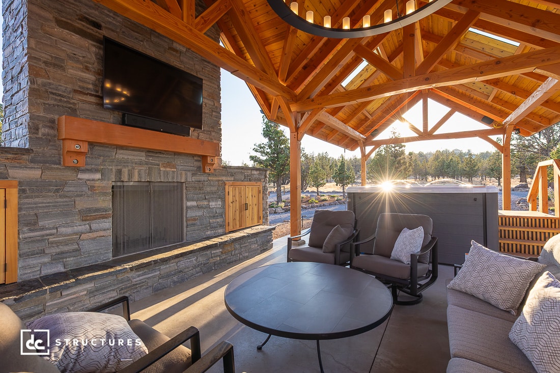 A cozy outdoor patio with stone fireplace, mounted TV, cushioned chairs, round table, and wooden pergola roof. Sunlight streams in.