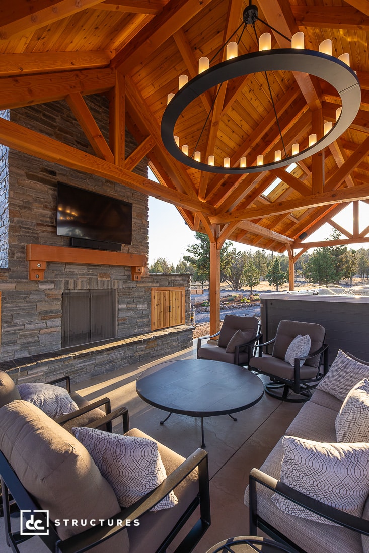A cozy outdoor seating area with cushioned chairs, a round table, stone fireplace, mounted TV, and wooden pergola roof. Trees in back.