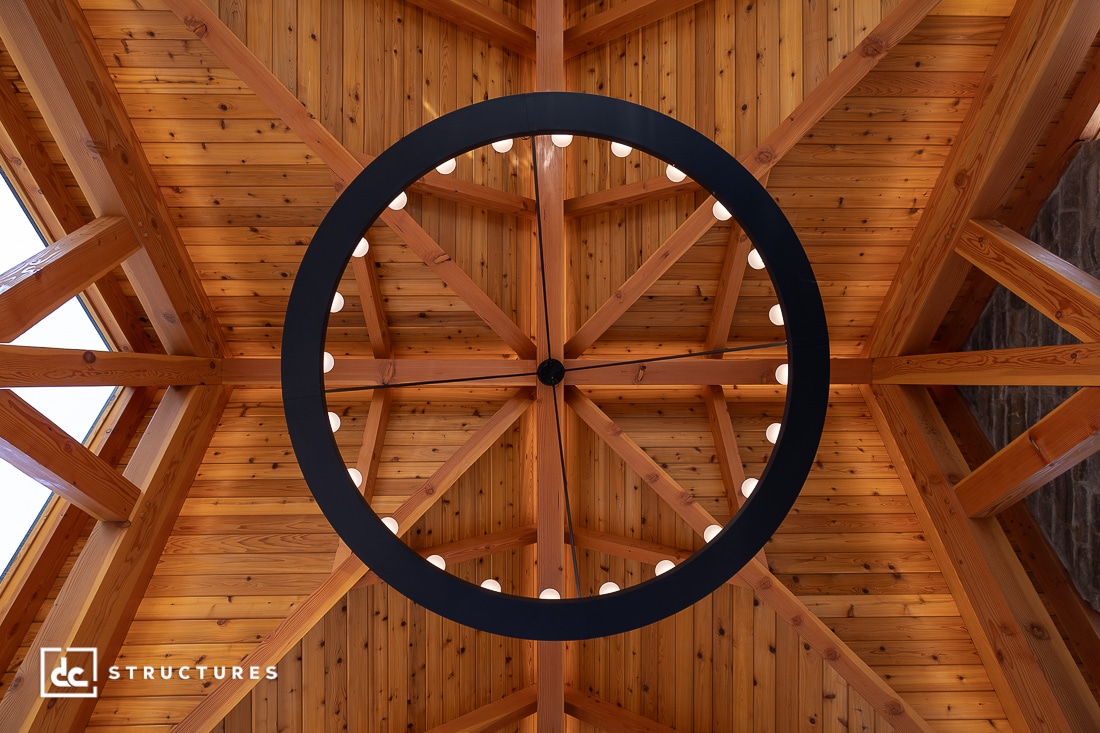 View of a wooden, vaulted ceiling with exposed beams and a circular black chandelier with lights, seen from directly below.