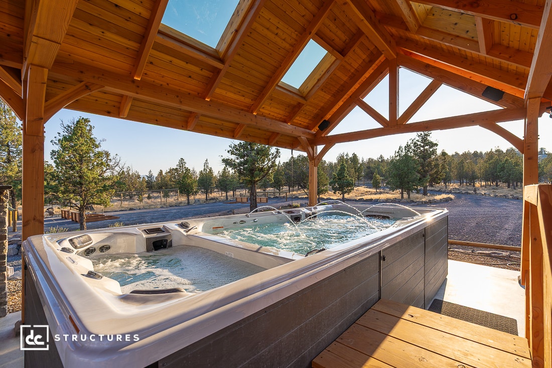 A large outdoor hot tub sits under a wooden pergola with skylights, overlooking trees and open sky in the bright sunlight.