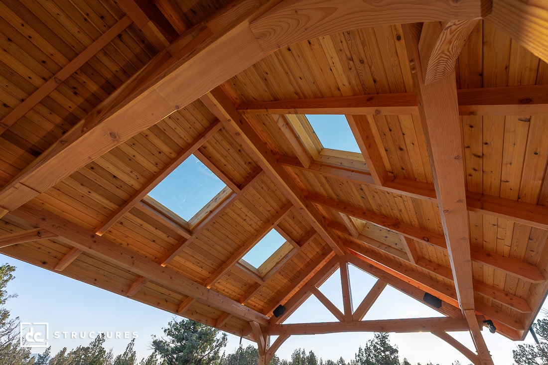 Wooden timber frame ceiling with exposed beams and multiple skylights, viewed from below. Sunlight streams in, trees outside.