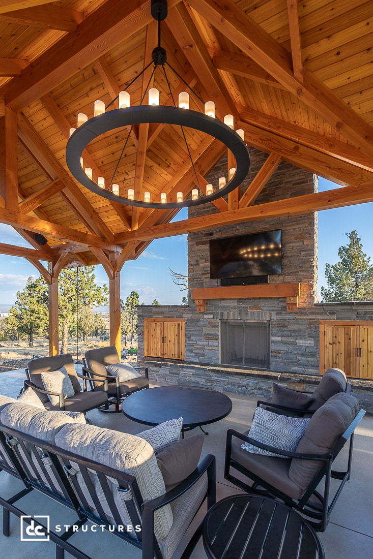 Covered outdoor patio with wooden ceiling, large round chandelier, stone fireplace, mounted TV, cushioned chairs, round table. Trees surround.